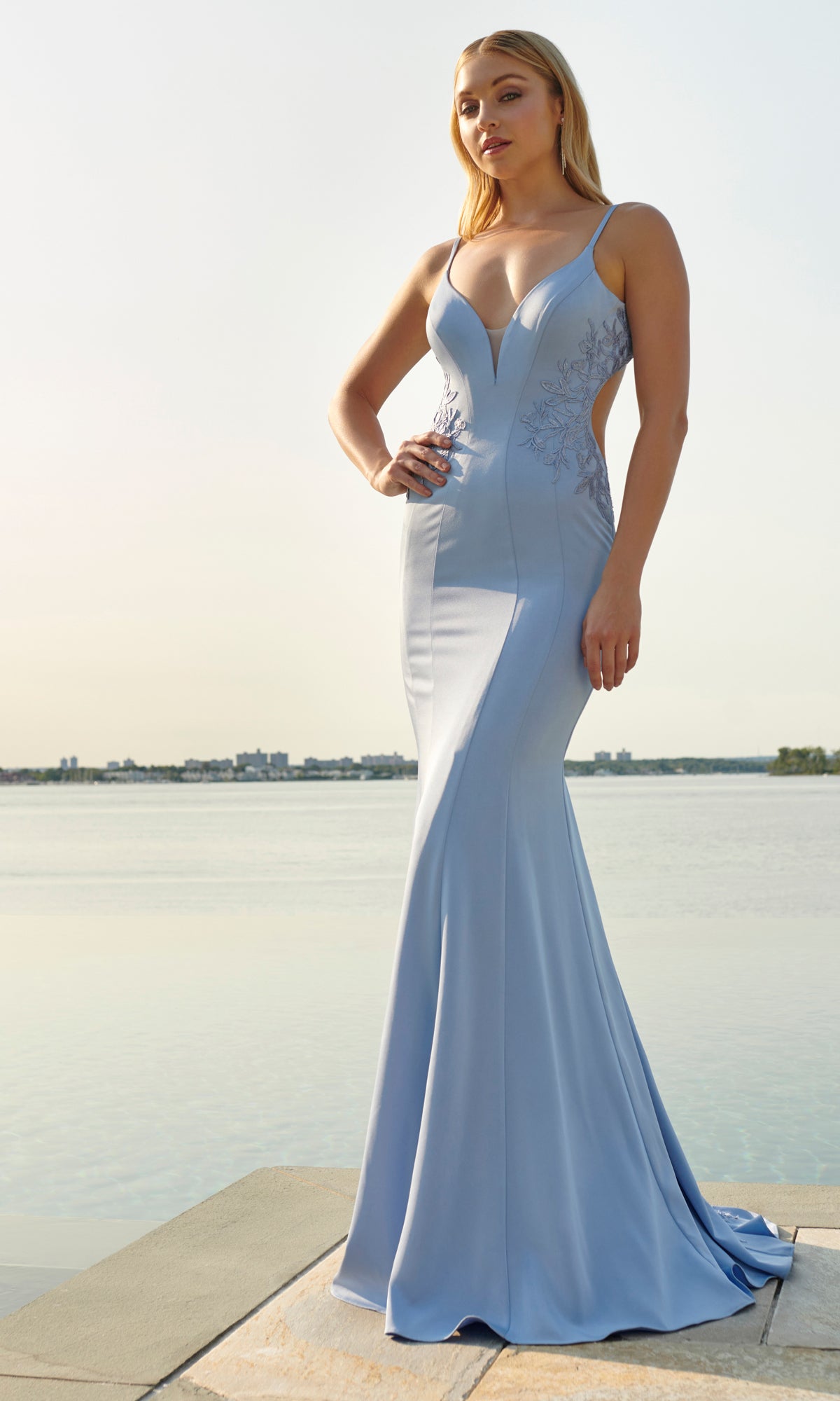 A woman stands by the waterfront on a sunny day, wearing the Faviana Long Satin Prom Dress with Side Cut Outs in light blue, featuring thin straps and elegant designer details.