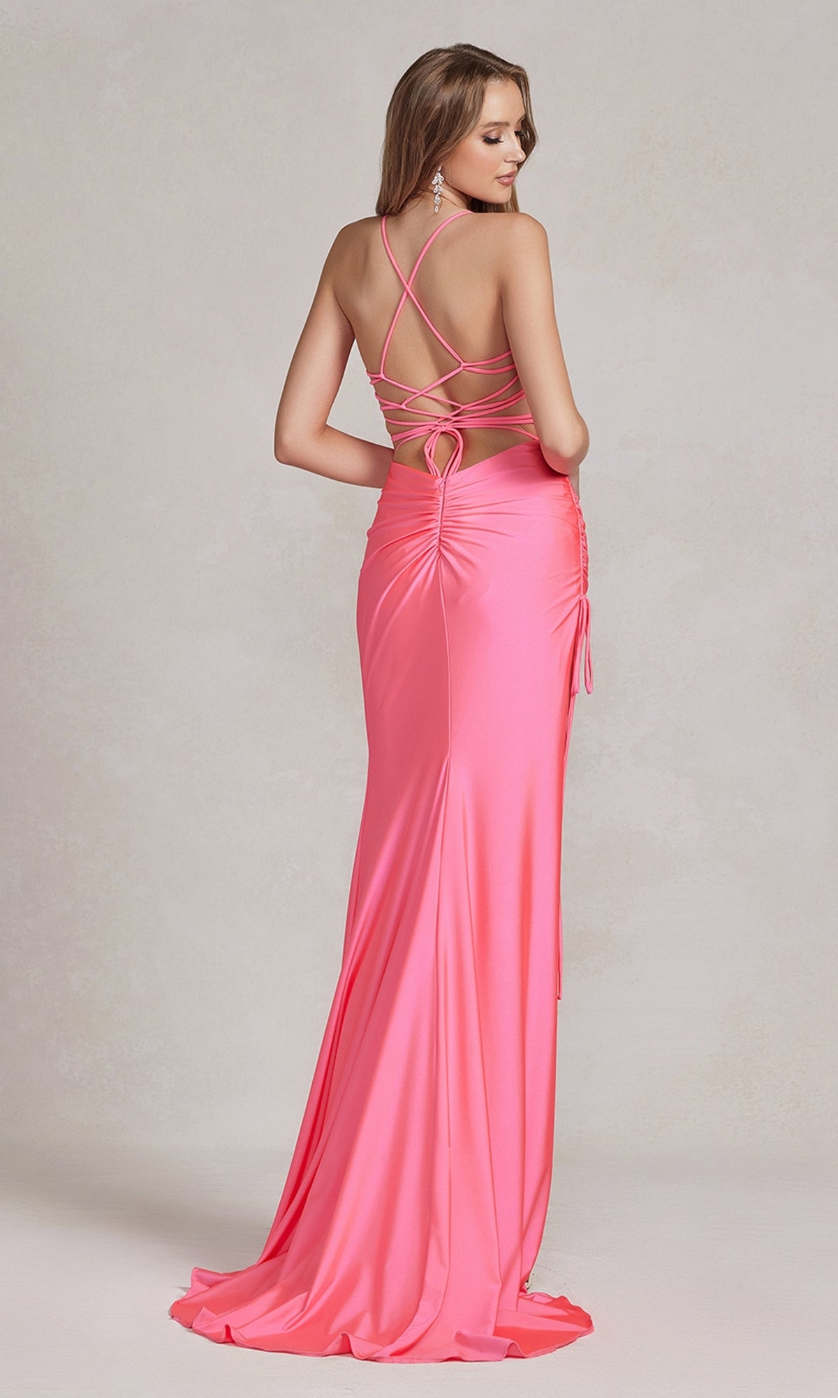 A woman wears the Strappy-Back Affordable Long Prom Dress, a floor-length bright pink gown with a fitted ruched back, crisscross open straps, and a heart-shaped cutout, standing against a light neutral background.