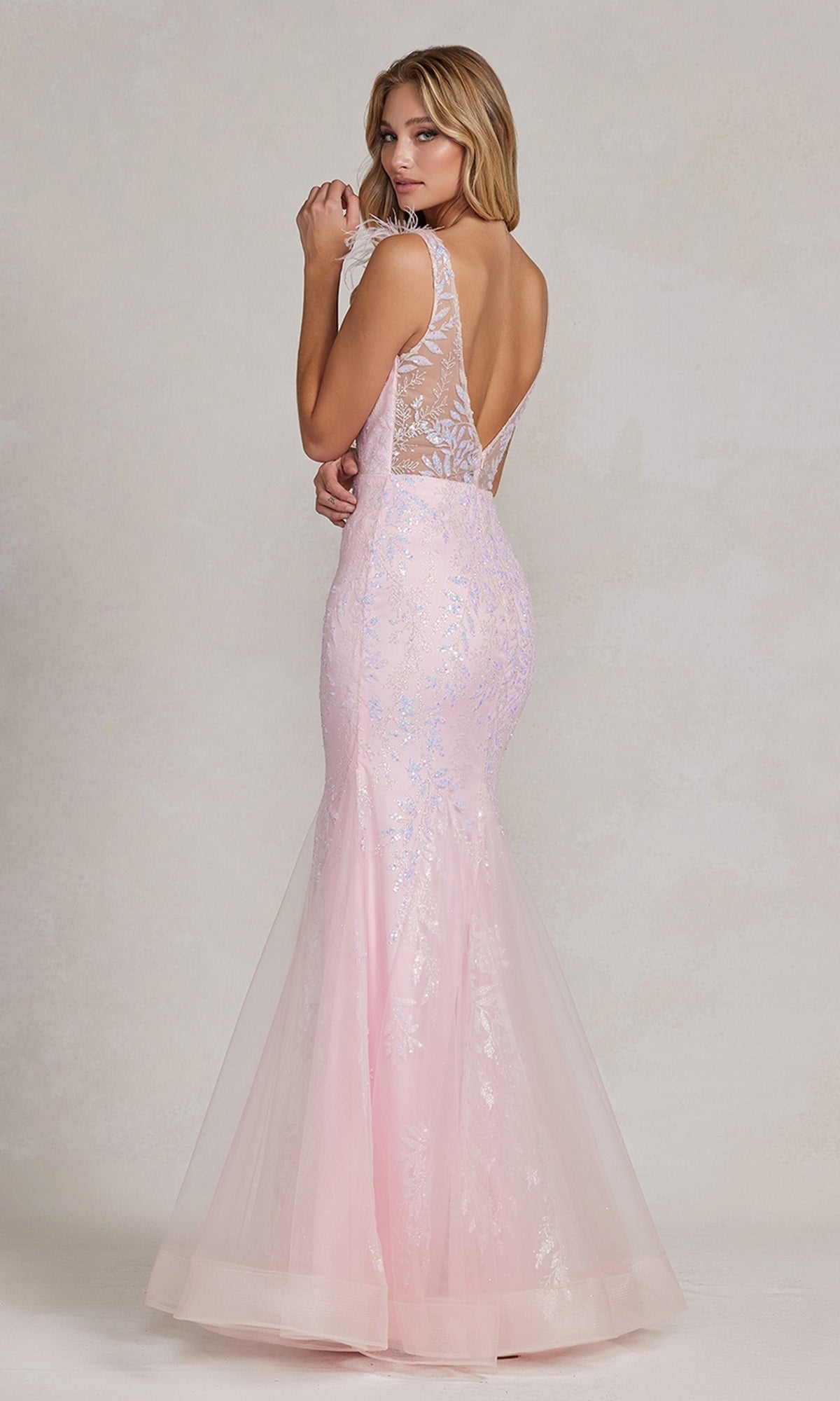 A woman, back slightly turned, wears the Feather-Strap Long Prom Dress: Nox Anabel T1208—a light pink, floor-length glitter mermaid style with a sheer lace back and subtle shimmer.