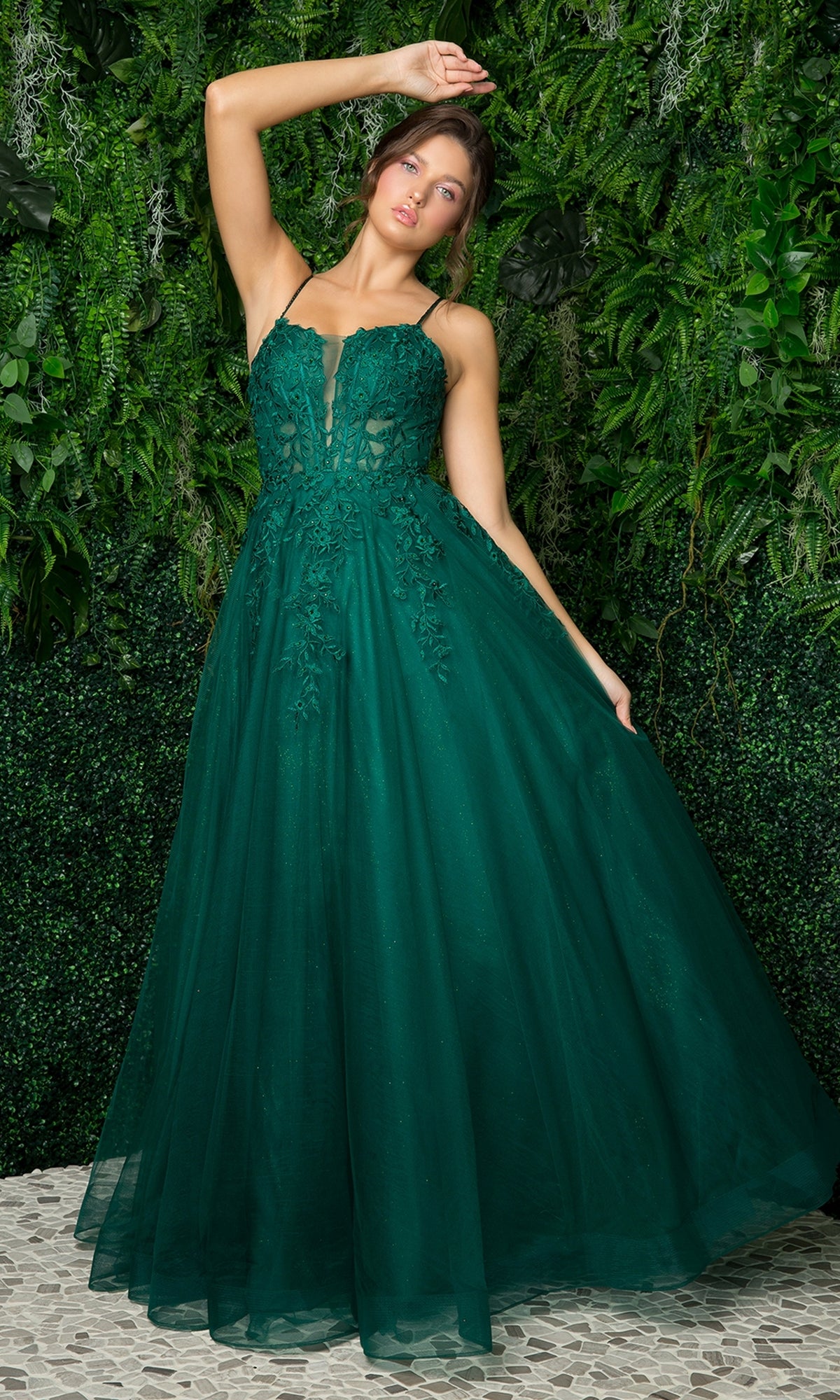 A woman in a Long Embroidered Sheer-Corset Prom Ball Gown—sleeveless, deep green, with a floor-length tulle skirt and floral details—stands before a leafy green backdrop.