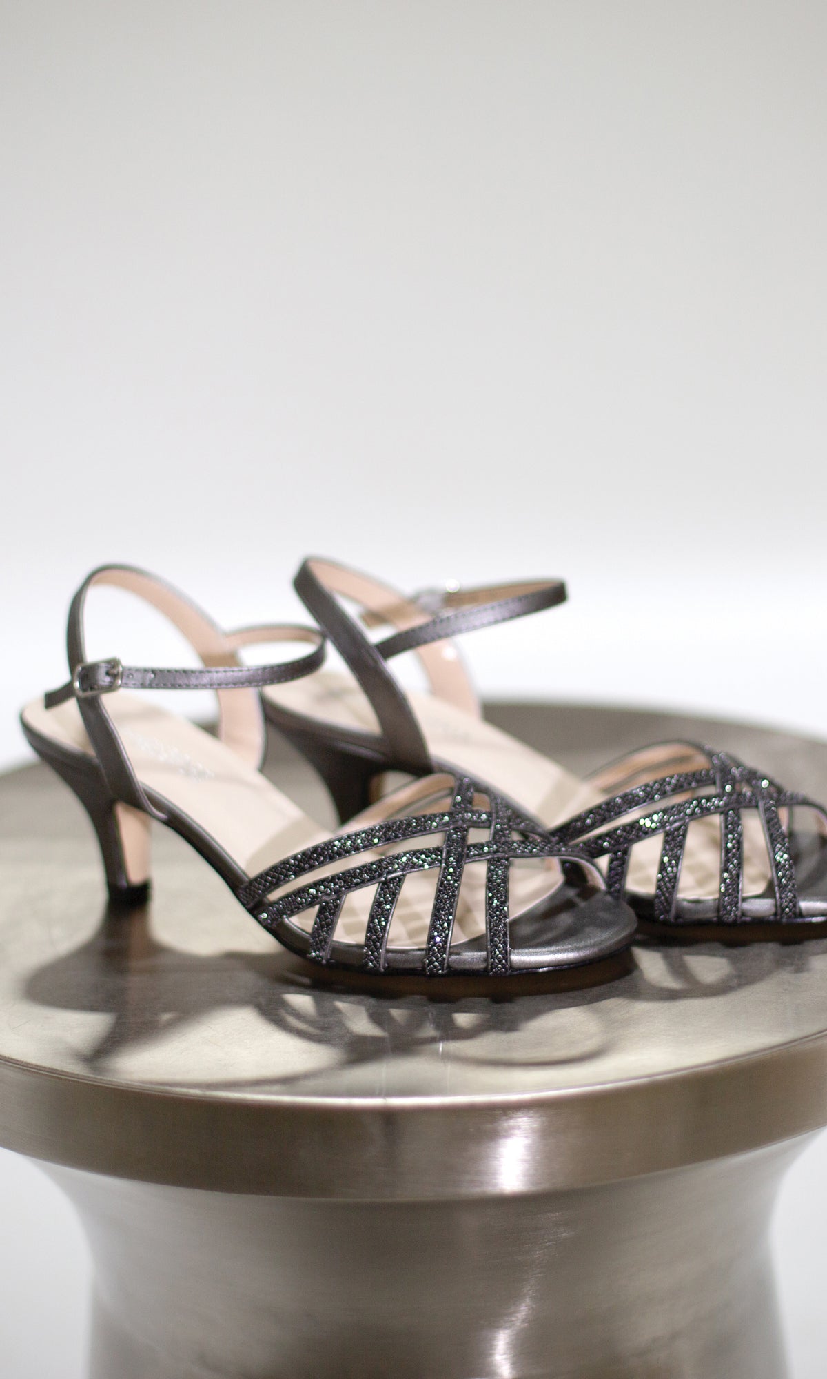 The Amara Pewter Open Toe Prom Shoes 4485, featuring stylish strappy details and a low heel, are elegantly displayed on a metallic round table, making them the perfect choice for prom.