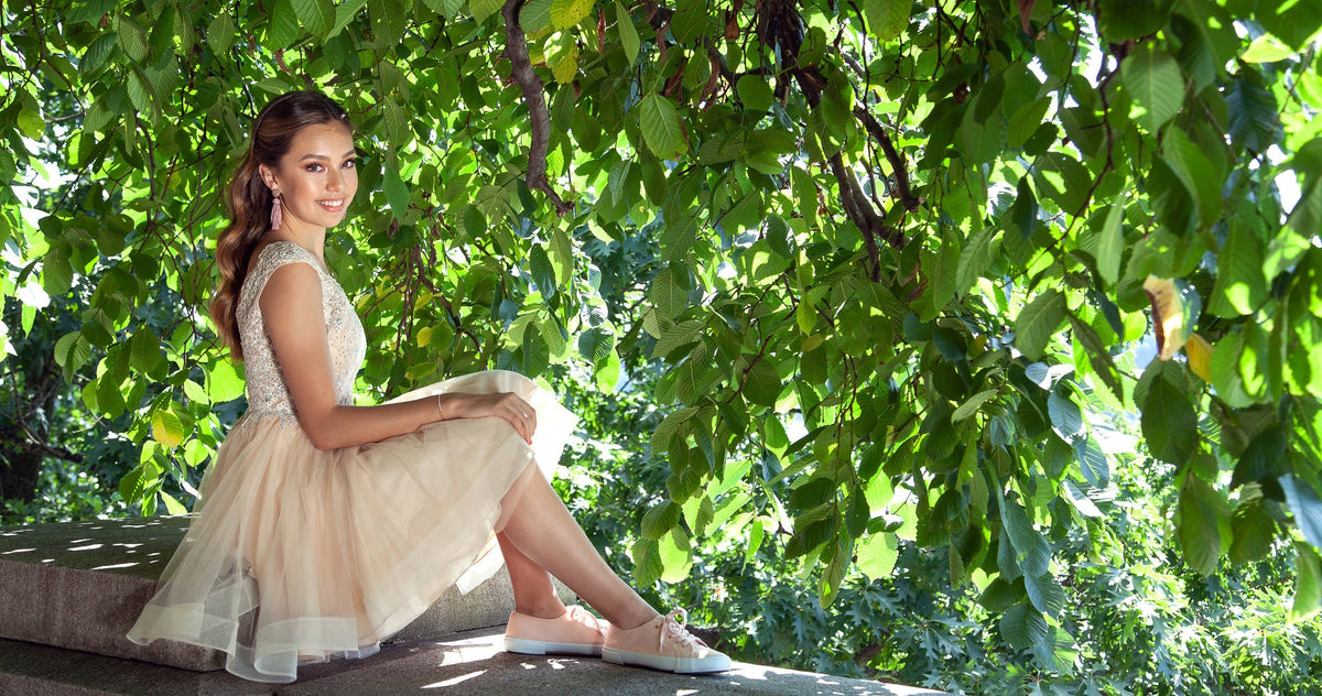 Teen girl in an ivory graduation dress sitting on a stone wall under a tree.