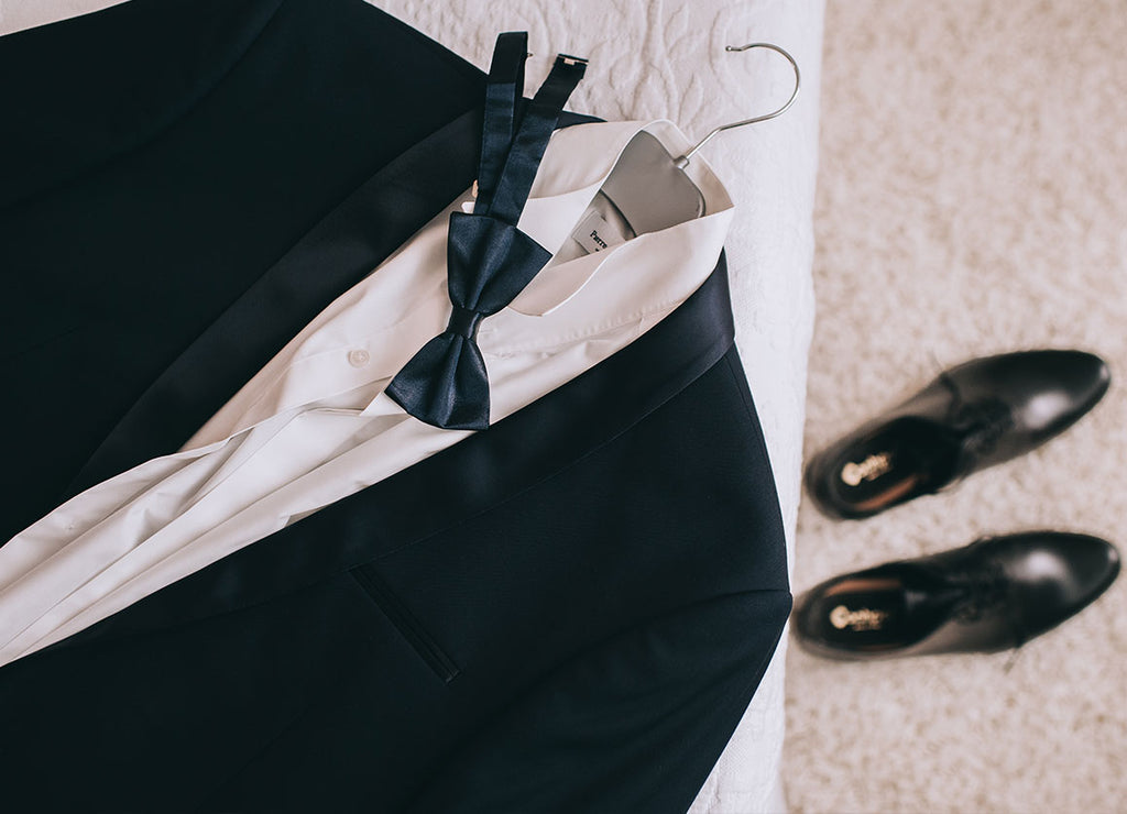A black bow tie rests on a white shirt and black tuxedo jacket on a hanger and black dress shoes are in the background.