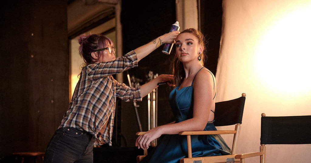 Wearing a homecoming dress, a teen girl sits tall in a director's chair while her hair is styled.