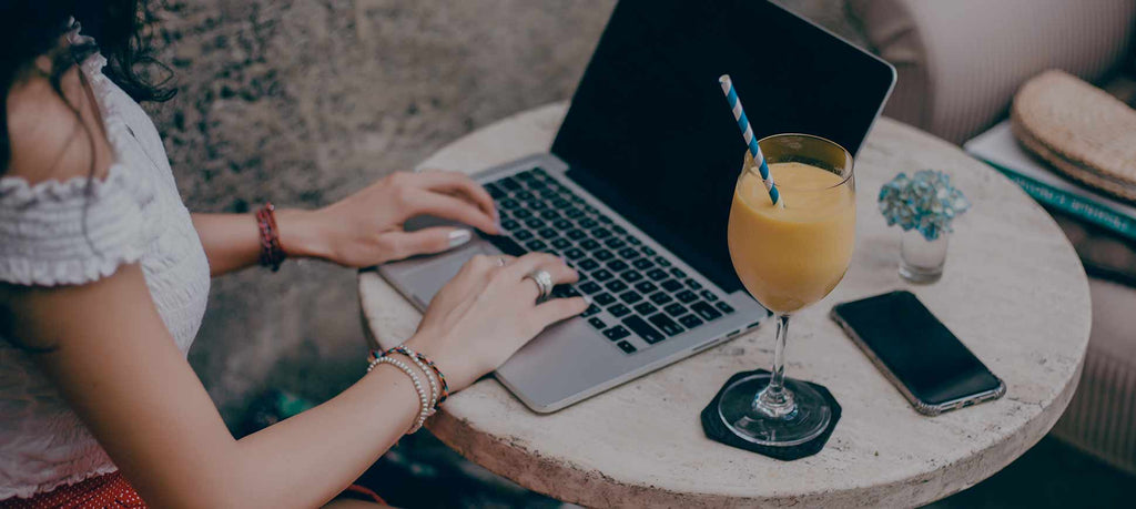 Close up of a girl's hands typing on her open laptop next to her phone and a wine glass with orange juice and a straw.