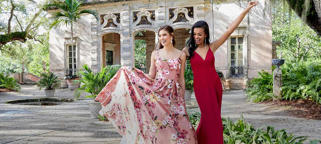 Two teen girls, one holding out the skirt of her floral prom dress and the other waving her arm in a red prom dress.