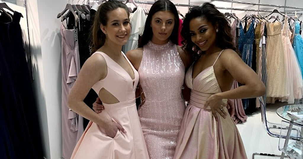 Three teens trying on homecoming dresses in front of a dress rack.