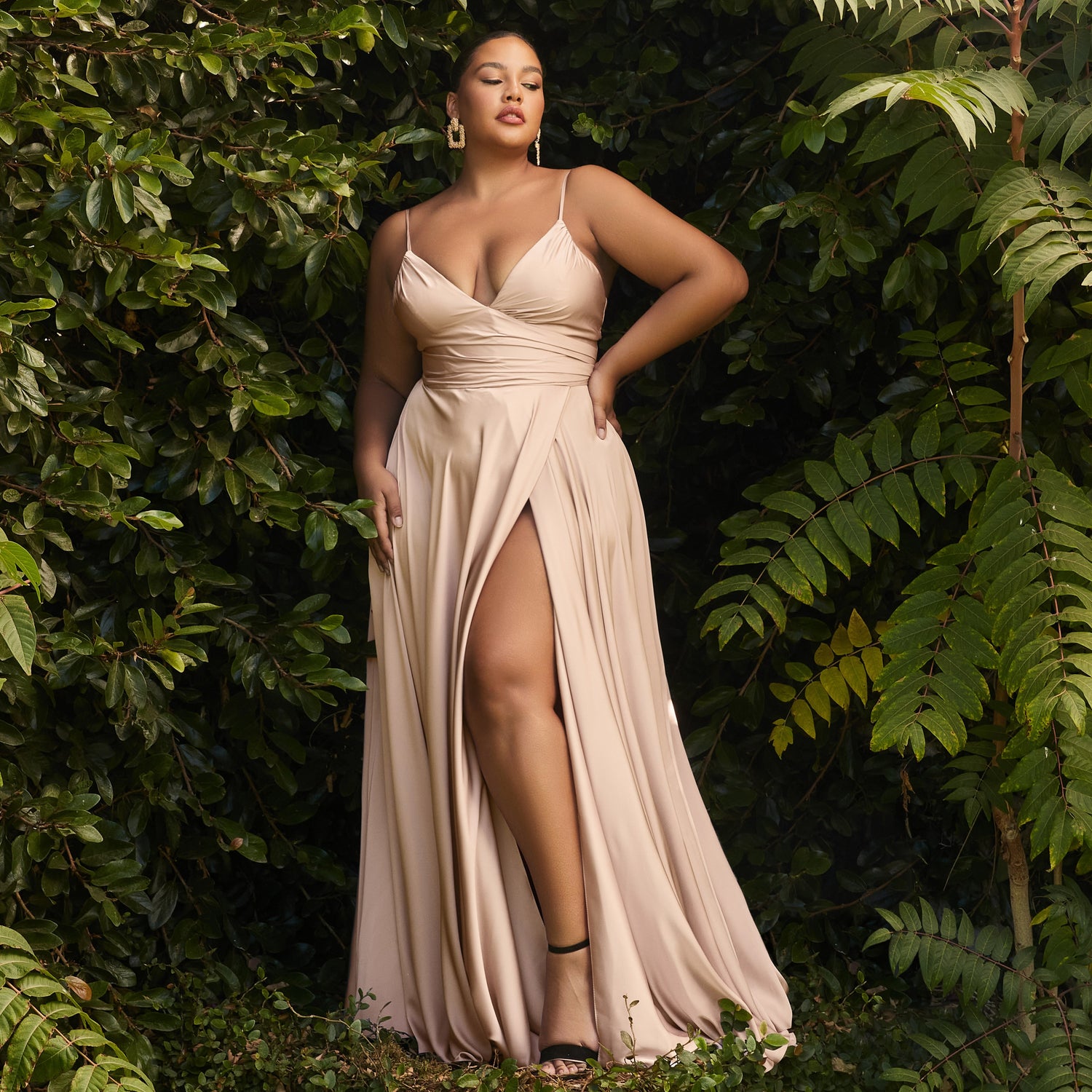 Image of a woman wearing a long plus-size nude colored prom dress standing outside surrounded by trees.