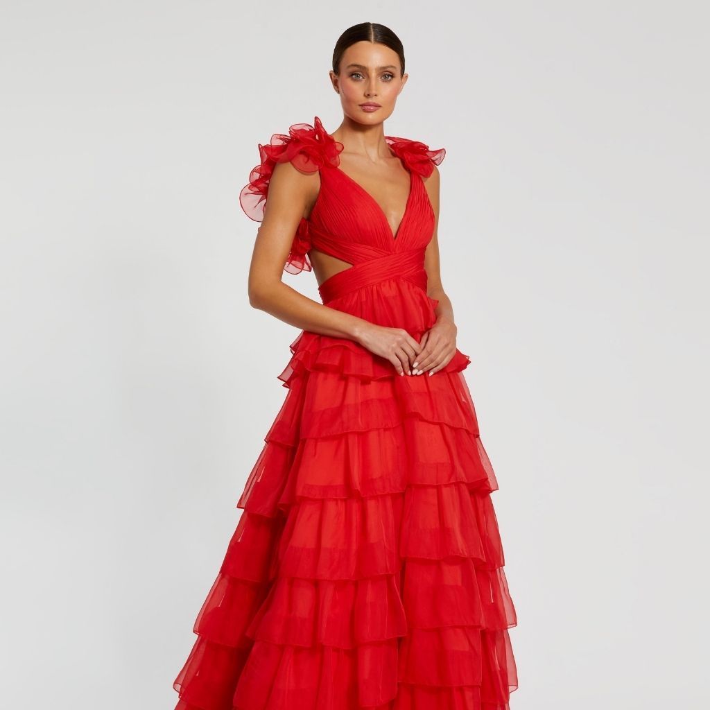 Against a white backdrop, a woman poses in a red military ball dress with ruffled straps and tiered skirt.