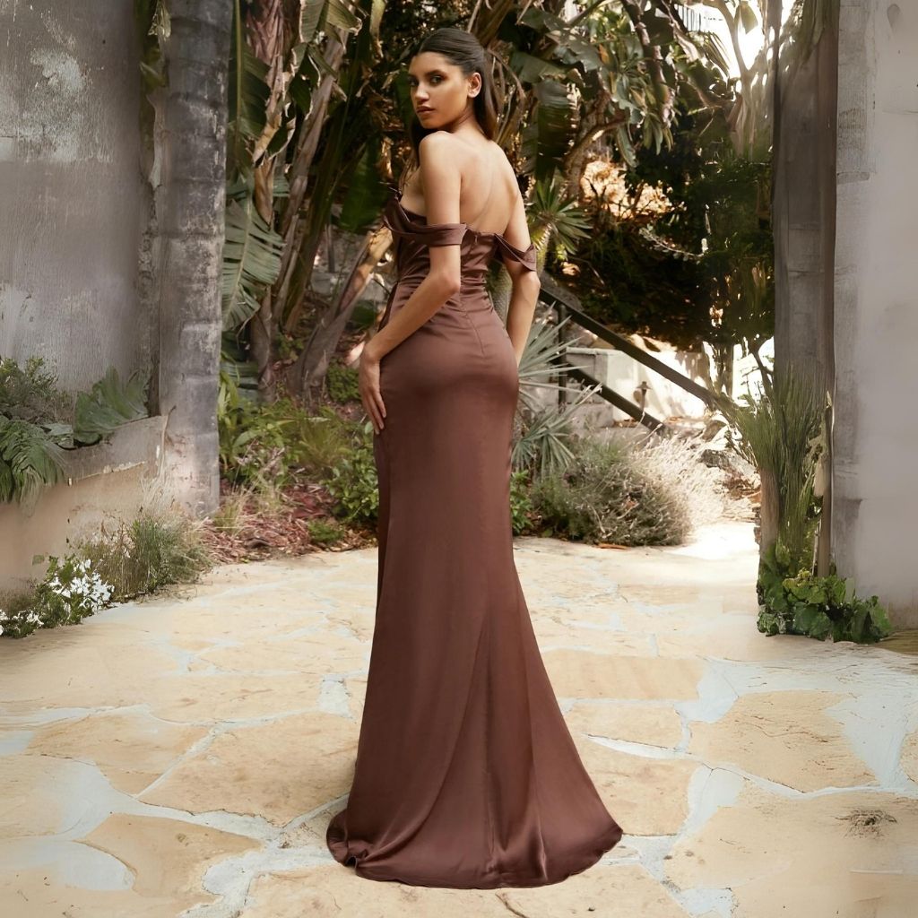 Wearing an off-the-shoulder brown prom dress, a teen walks toward a forest backdrop and stops to look back at the viewer.