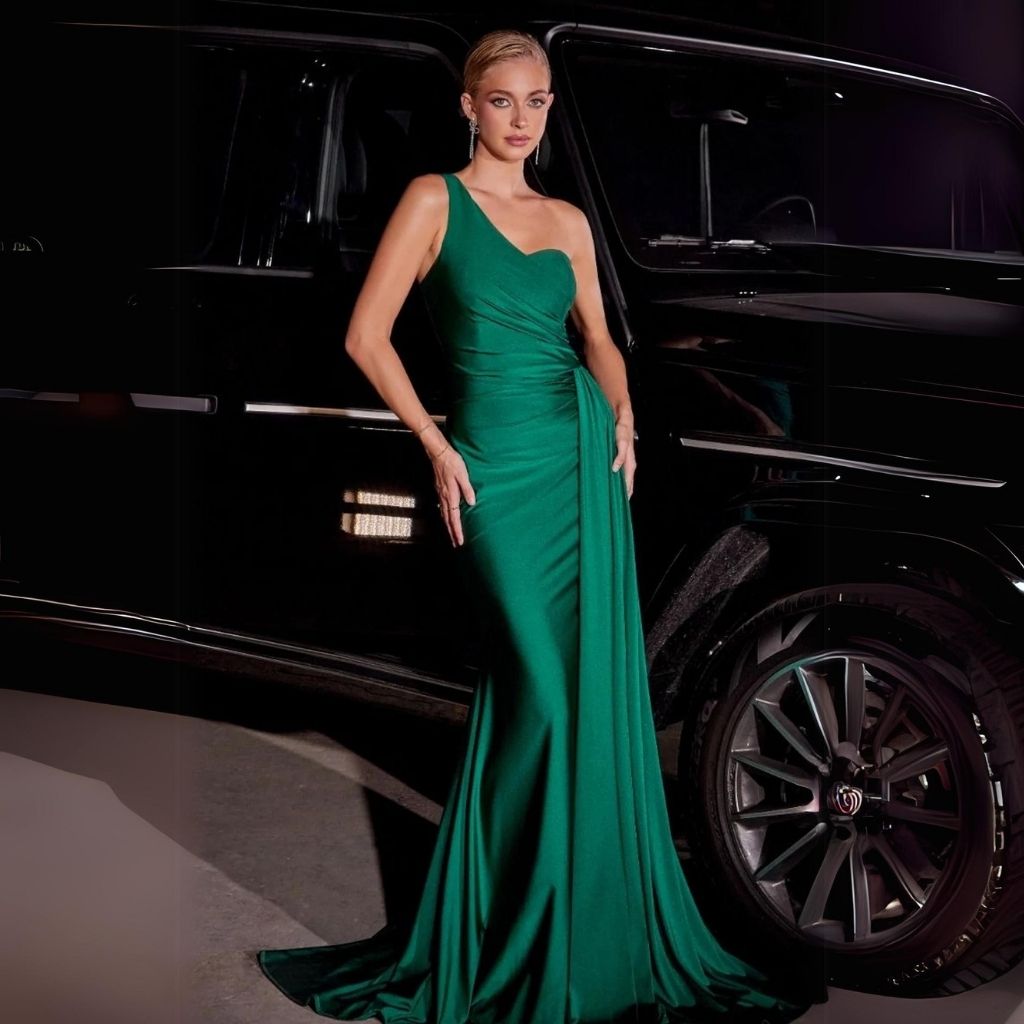 In a one-shoulder ruched classic green prom dress with a side sash, a teen girl stands beside a black car at night.