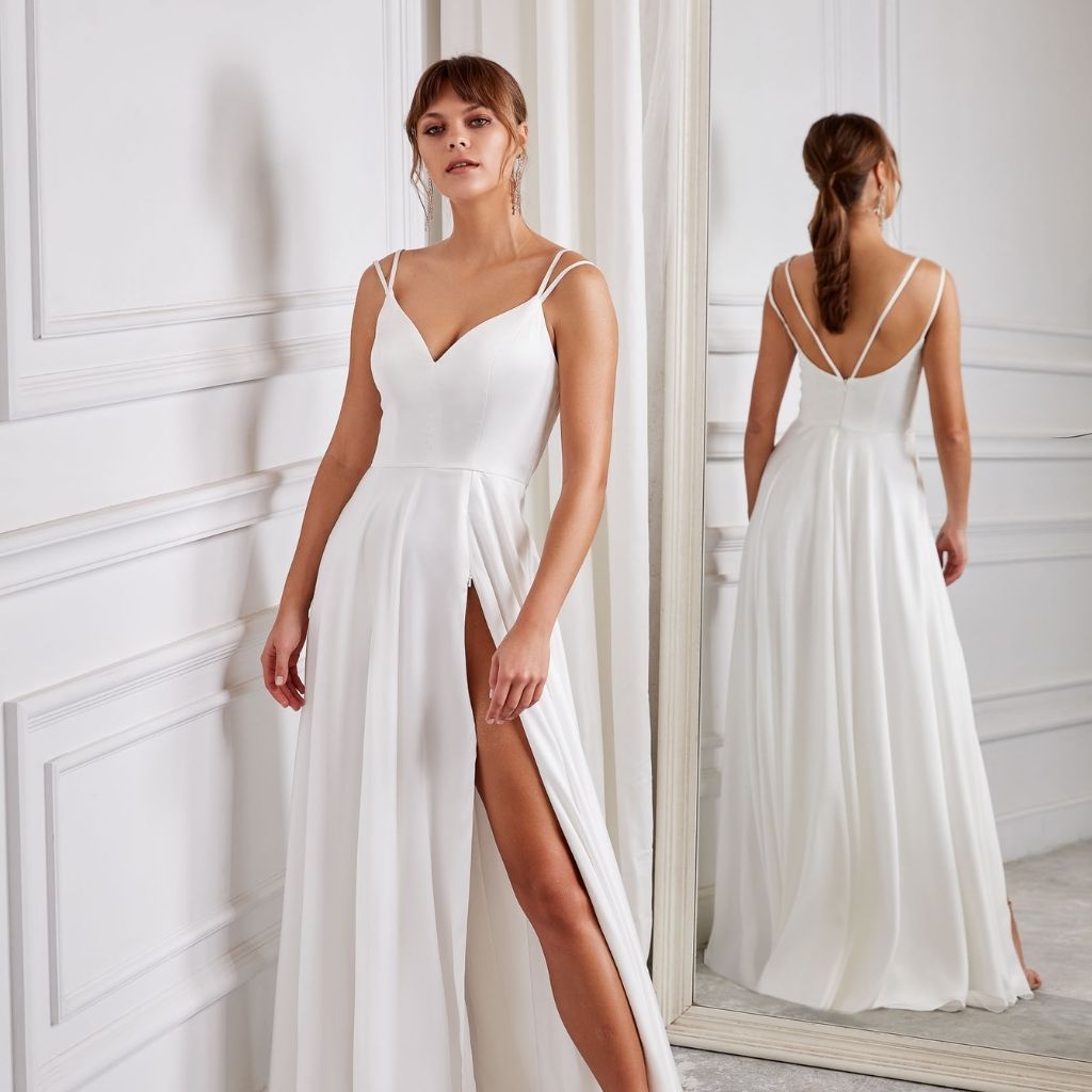 In a simple a-line white formal dress with double straps, a woman stands before a mirrored wall.