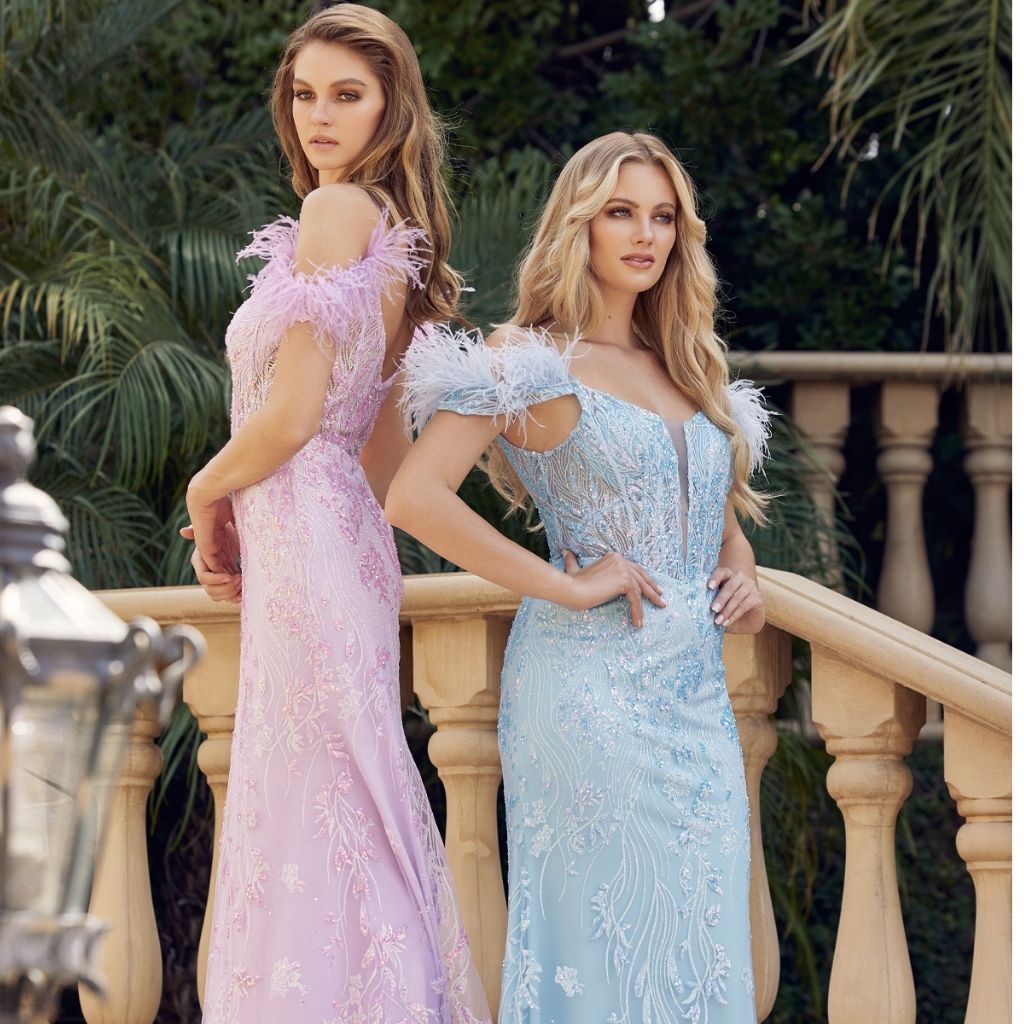 In a light purple and a light blue sheer corset prom dress by Juliet, two teen girls stand back to back by a stone railing.