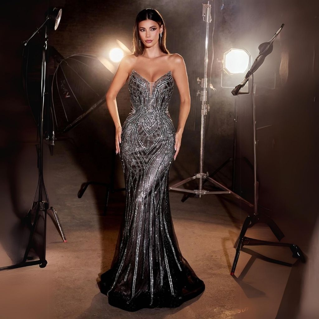 In a photo studio, a woman poses in a strapless long black fitted prom dress with a geometric silver sequin print.