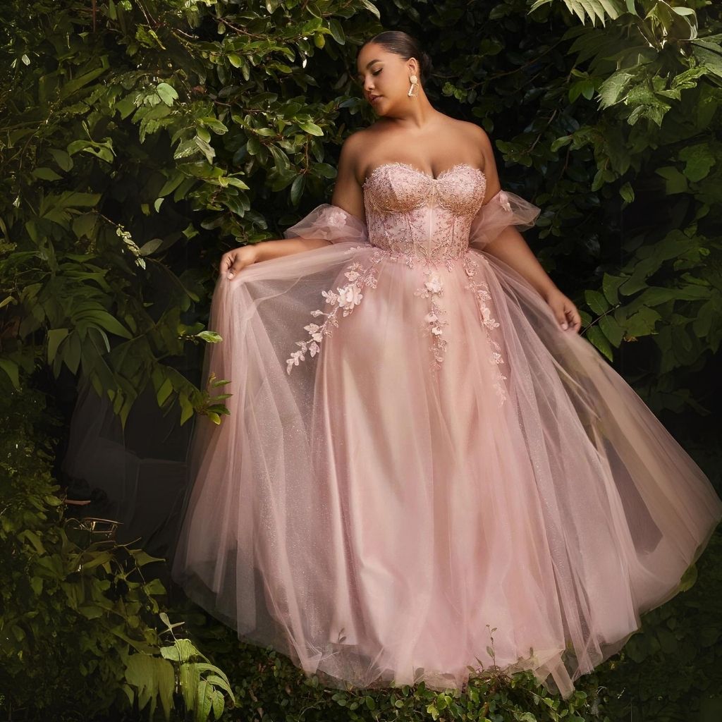 A curvy woman is standing among green leafy plants while wearing a strapless sweetheart plus-size prom dress.