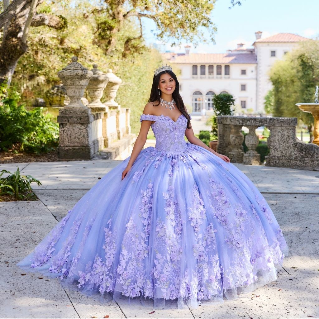 In a lilac purple off-the-shoulder floral quinceanera dress from Princesa by Ariana Vara, a girl stands in a stone courtyard.