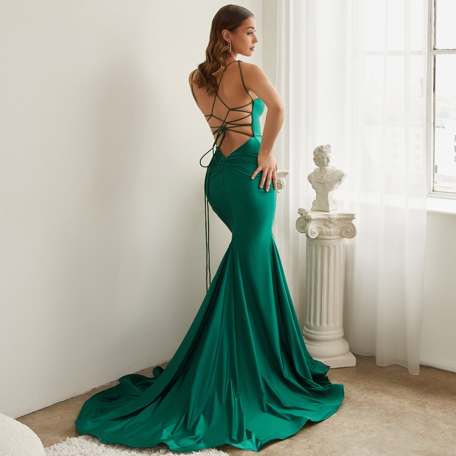 Image of woman standing looking out the window showing off her long emerald green open back prom dress.