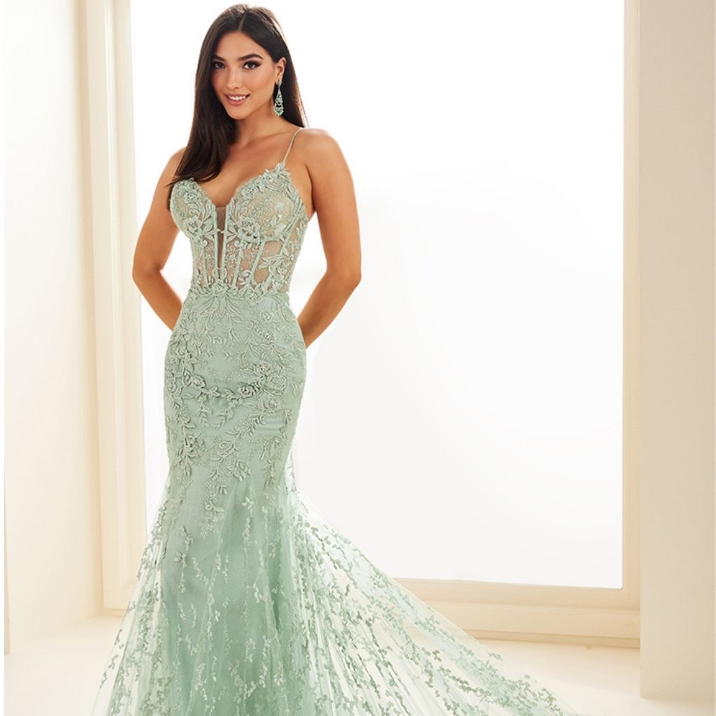 In front of a lit window, a teen girl stands wearing a pastel green lace prom dress with sheer corset.