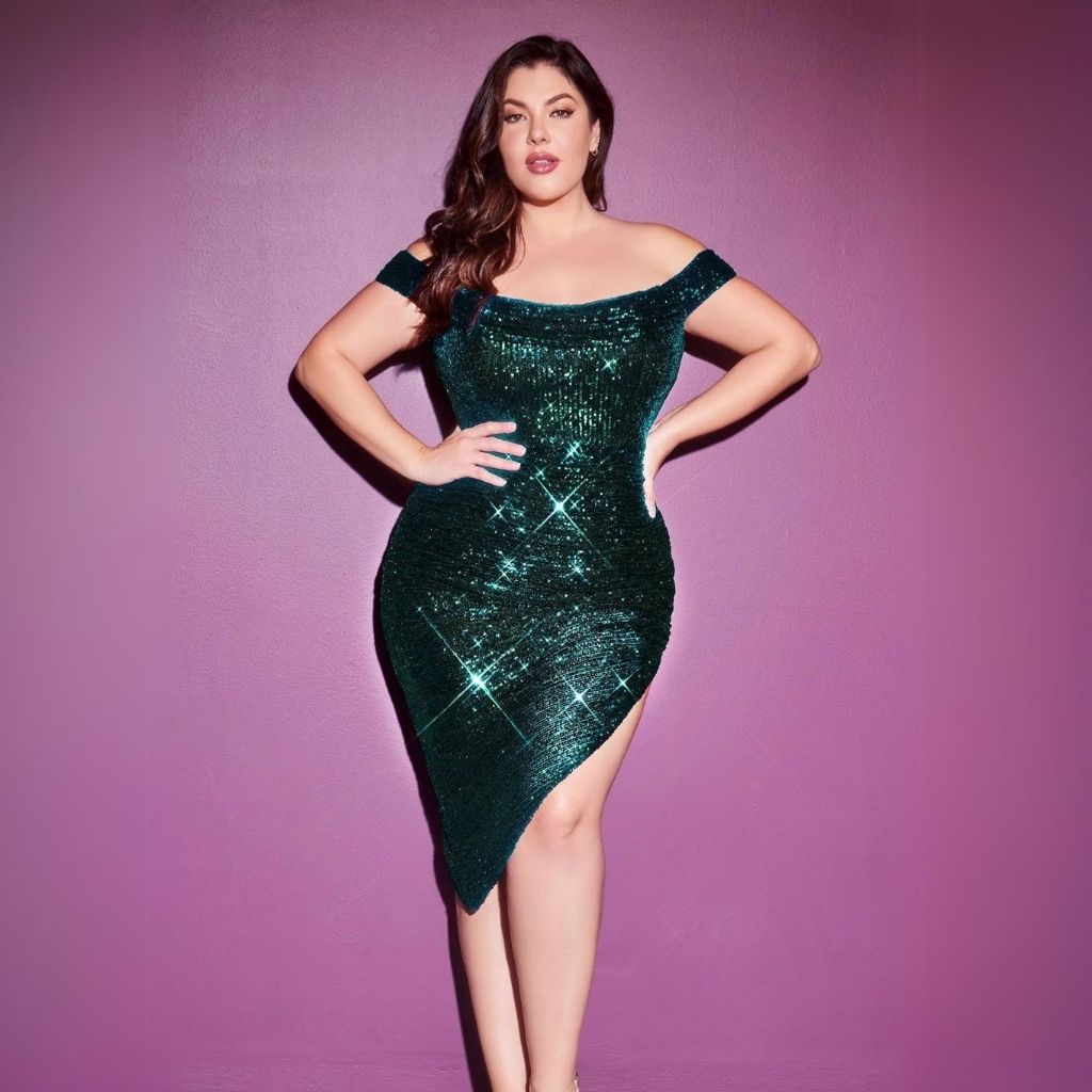 Against a purple backdrop, a teen girl wears an emerald green plus-size homecoming dress with side slit.