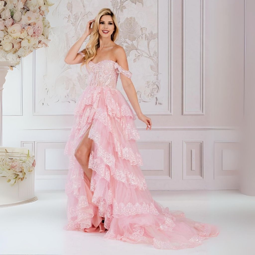 Against a decorative white backdrop, a woman poses in a pink lace prom dress with a tiered ruffled skirt and side slit.