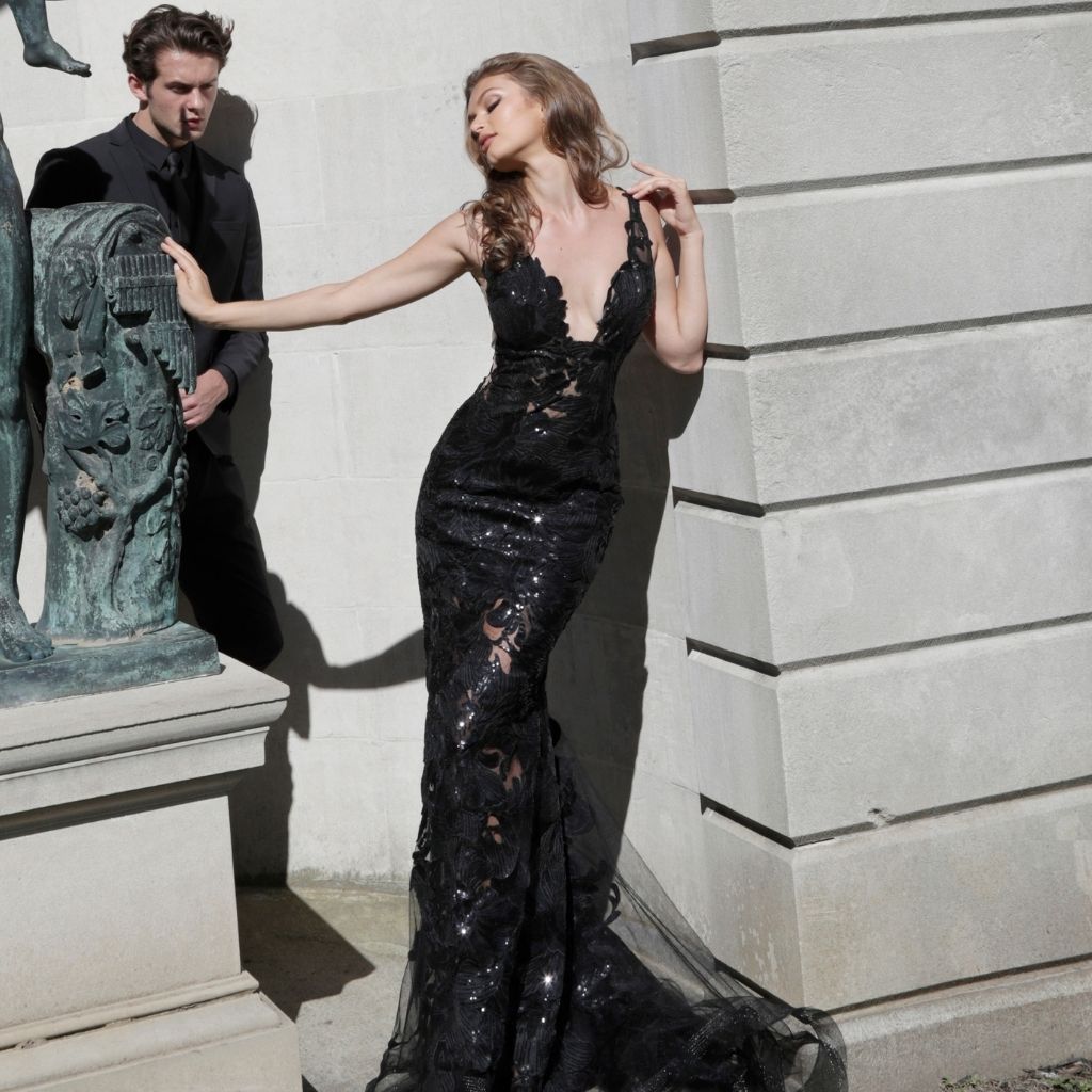 In a black sequin lace Jovani formal dress with plunging neckline, a woman leans against a stone wall.