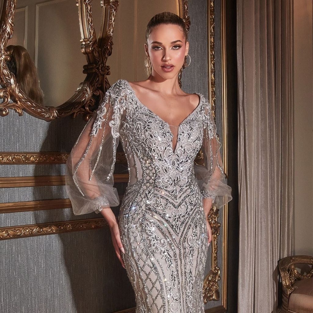 In a beaded silver formal dress with sheer long sleeves, a woman stands next to an ornate mirror.