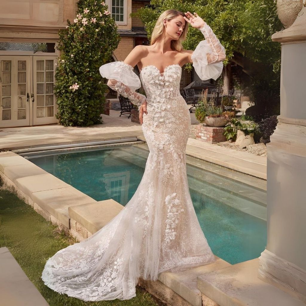 In front of an ornate pool, a bride poses in a strapless floral lace wedding dress with removable sheer puff sleeves.