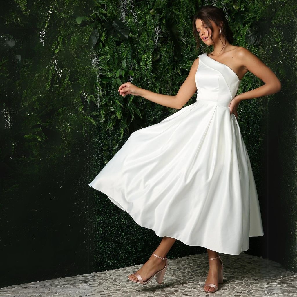 In a knee-length one-shoulder simple white reception dress, a bride walks along a green leafy wall.