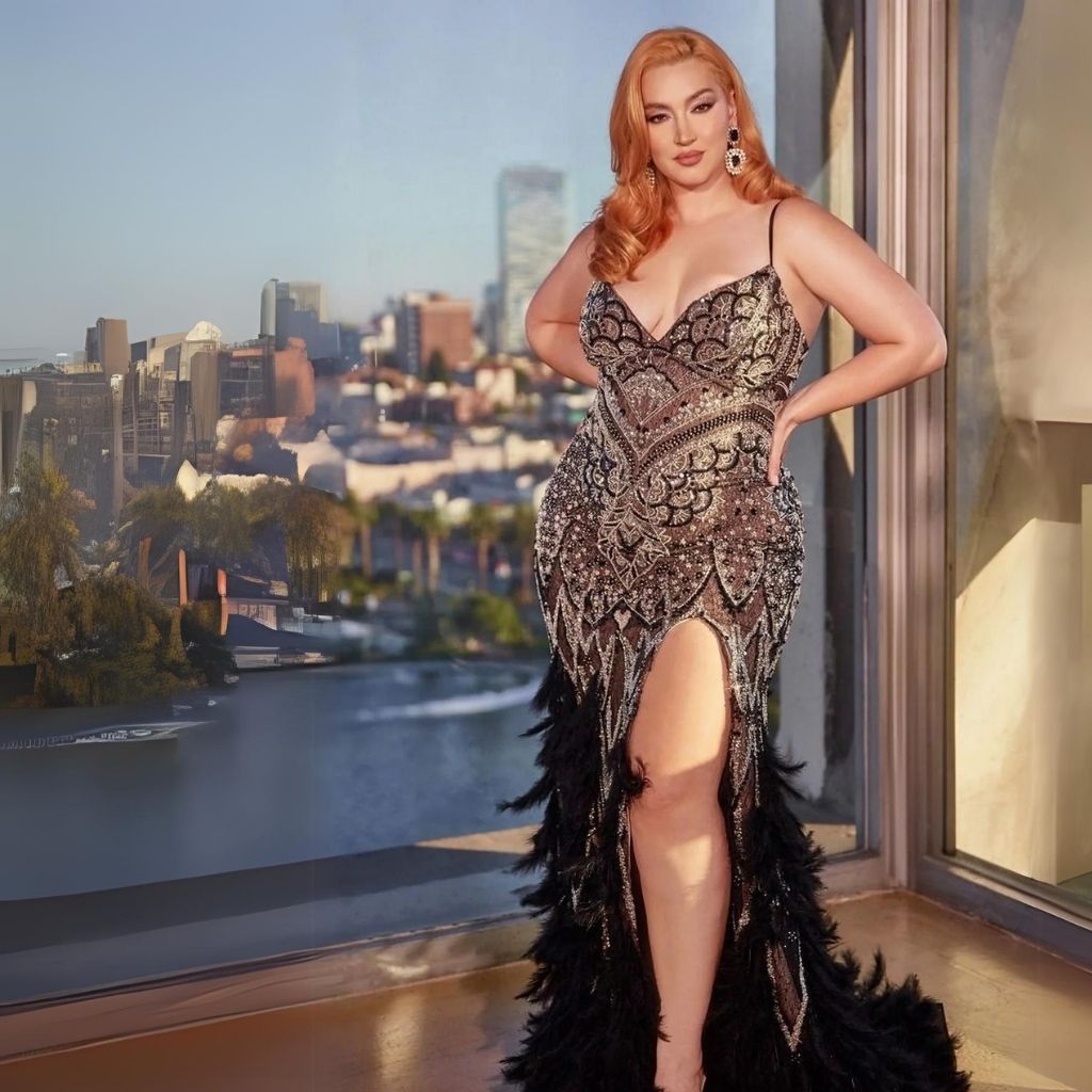 In a beaded plus-size formal dress with feather details, a woman stands in front of a floor-to-ceiling view of a city.