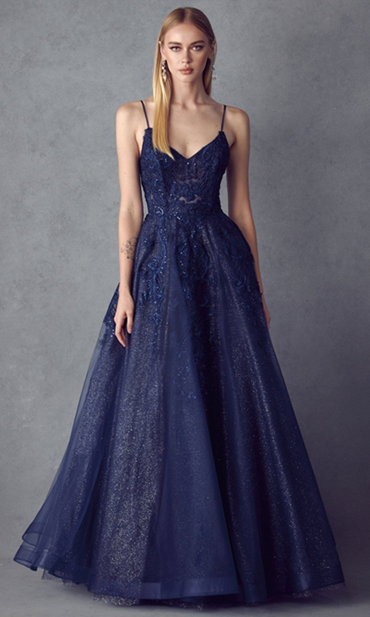 A woman wears the Embroidered Glitter-Tulle Long Prom Ball Gown 251, a sleeveless deep blue dress with a fitted bodice, flared skirt, lace, and glitter details, standing against a plain gray background.
