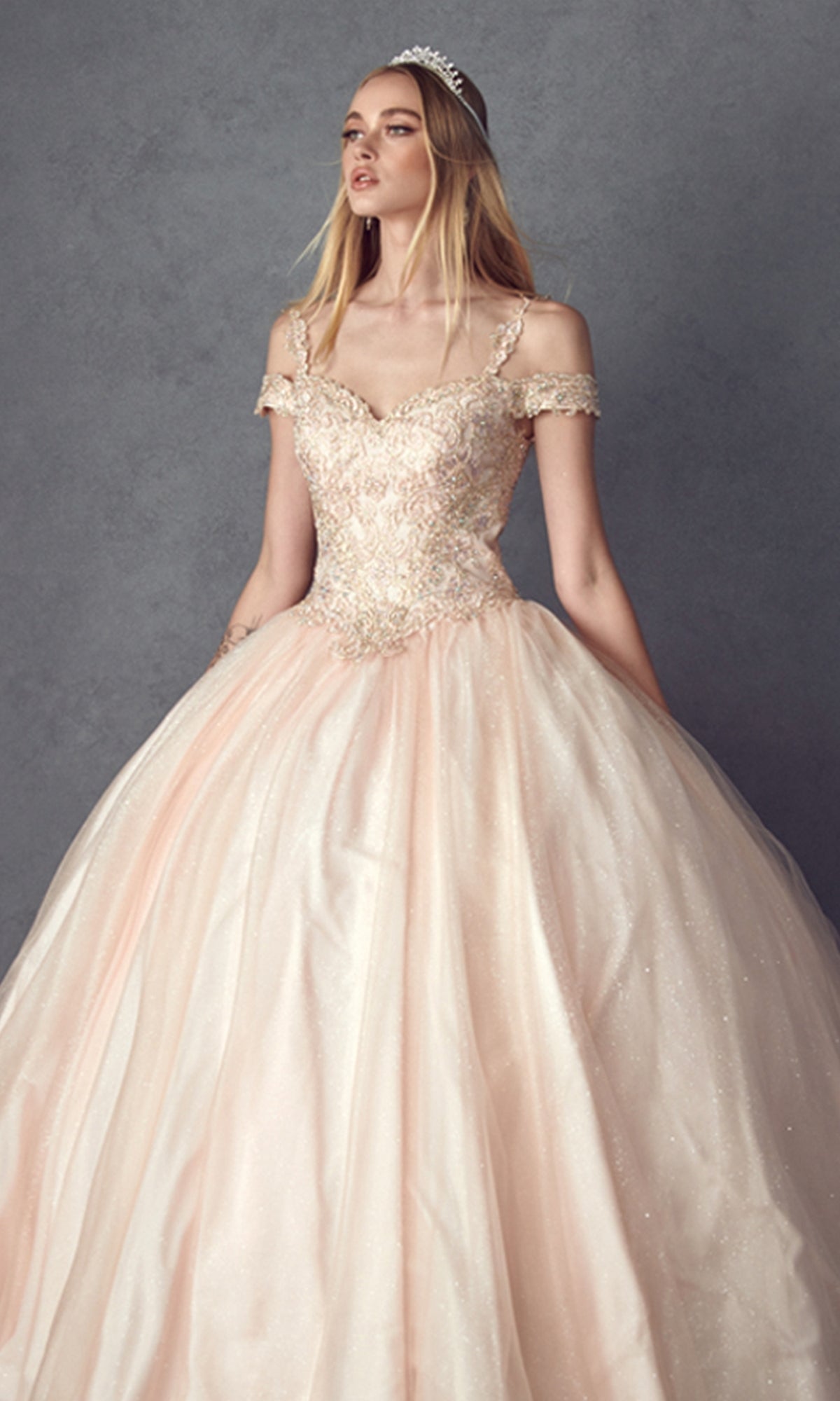 A woman wearing the Juliet 1430 Cold-Shoulder Glitter Quinceanera Dress with an off-the-shoulder neckline and full skirt, paired with a tiara, stands in front of a gray background.