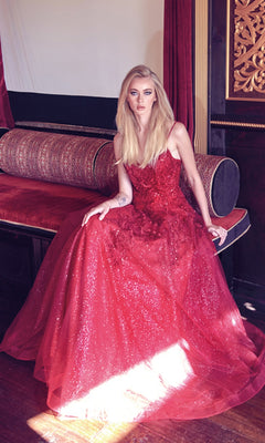 A woman with long blonde hair sits on a red and gold divan, wearing the Embroidered Glitter-Tulle Long Prom Ball Gown 251—a floor-length, sleeveless formal dress with floral details in shimmering red.