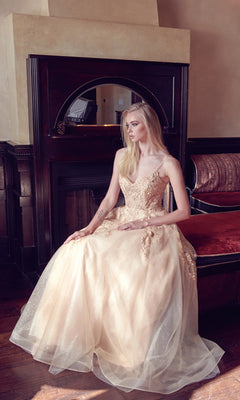 A woman wearing the Embroidered Glitter-Tulle Long Prom Ball Gown 251 sits on a red cushioned bench in front of a dark wooden fireplace.