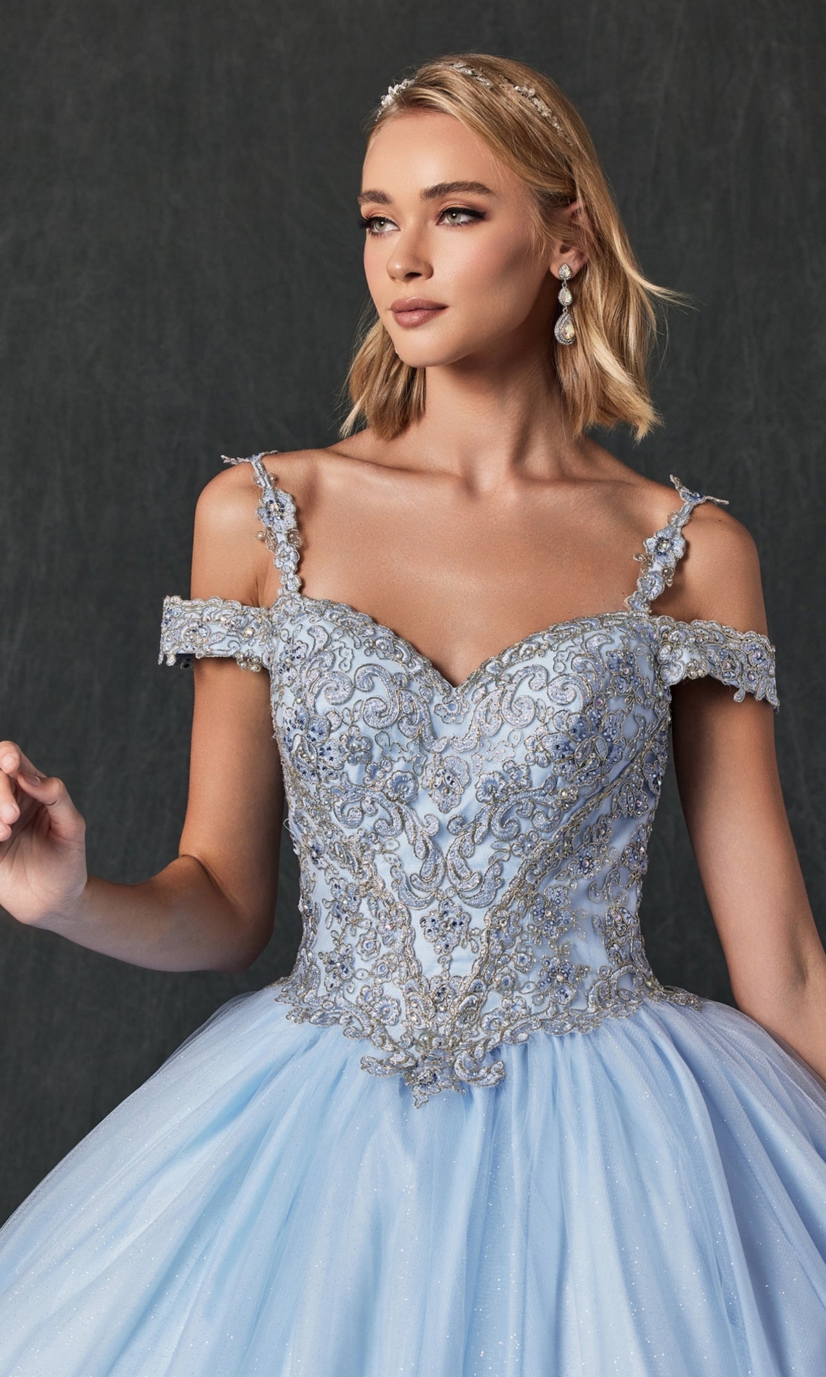 A woman wears the Juliet 1430 Cold-Shoulder Glitter Quinceanera Dress in light blue with silver embroidery, posing against a dark background.