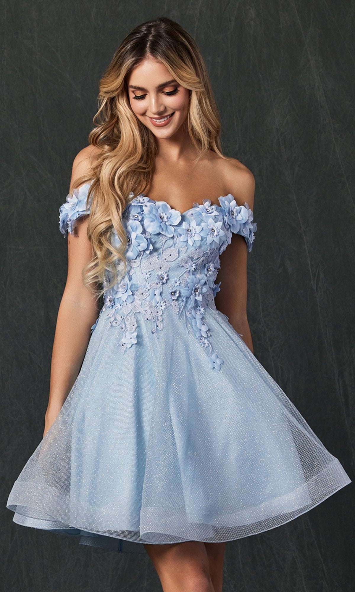 A smiling woman wearing the Floral Off-the-Shoulder Short Homecoming Dress 861, featuring 3-D flowers and a tulle skirt, stands against a dark textured background and looks down.