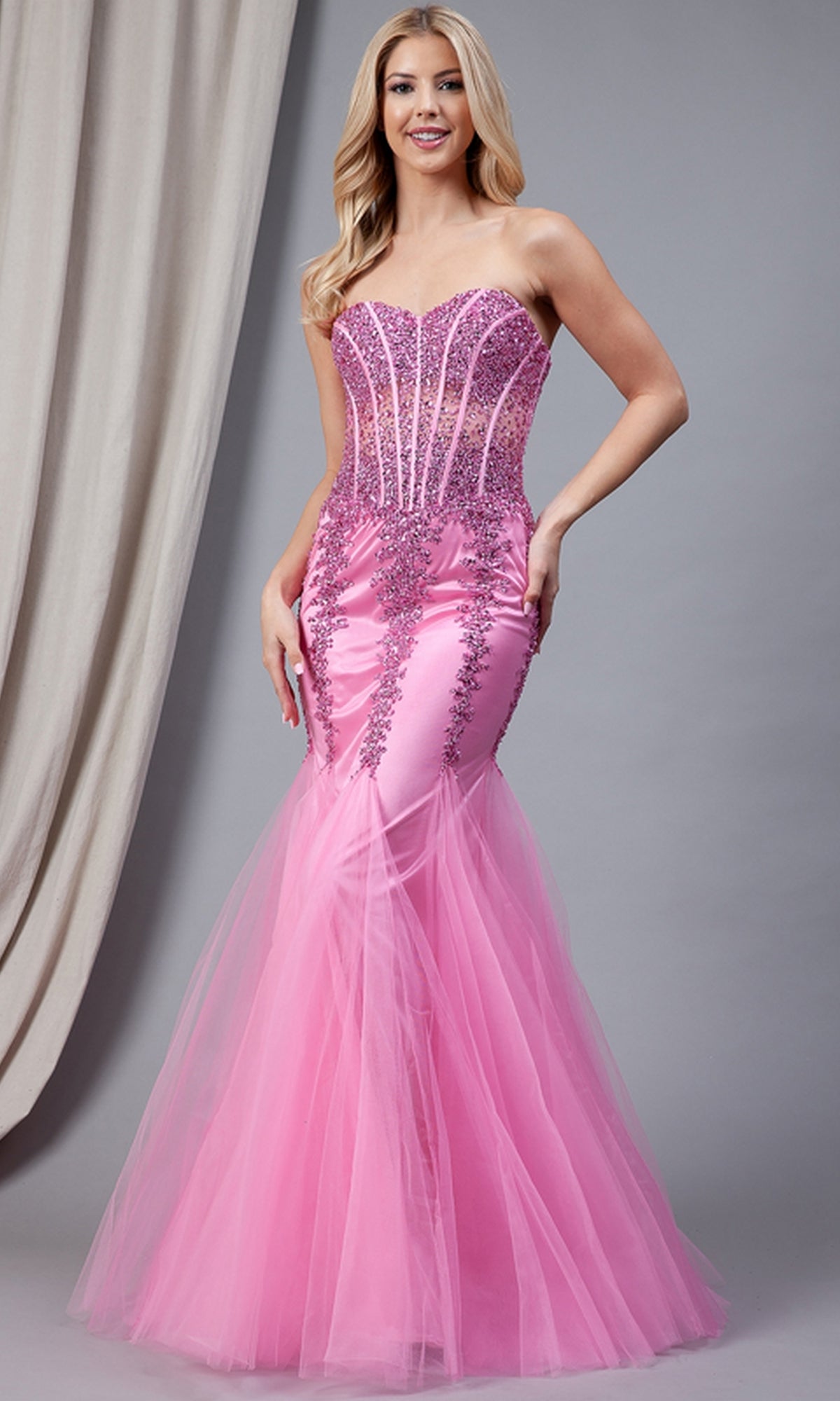 A woman wears the Sheer-Corset Strapless Mermaid Prom Dress 774, featuring a jeweled corset bodice, sequin accents, and a tulle skirt, standing before a gray backdrop with beige drapery.