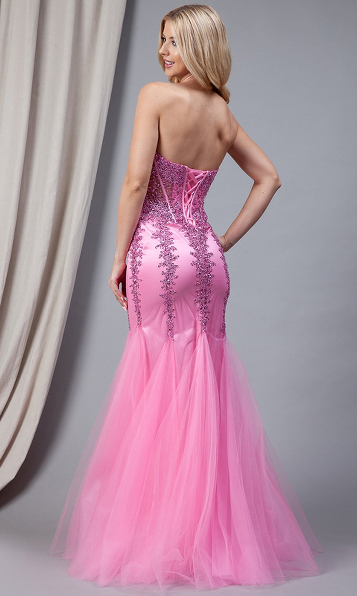 A woman wearing the Sheer-Corset Strapless Mermaid Prom Dress 774 with a jeweled corset bodice and lace details stands with her back to the camera, its floor-length tulle skirt elegantly flowing behind her.