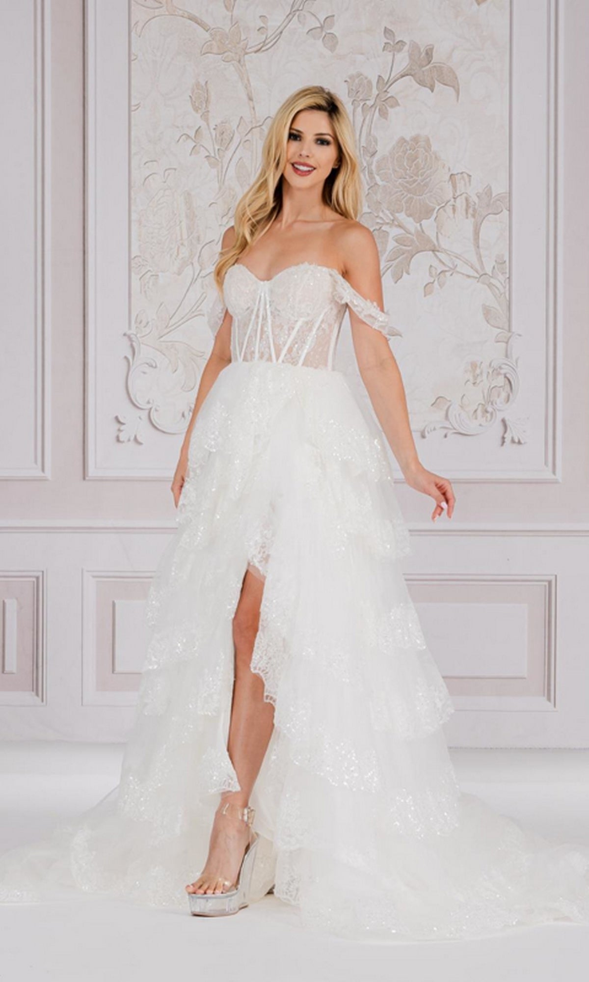 A woman poses elegantly indoors against a floral wall, wearing the Off-the-Shoulder Layered Long Ball Gown TM1012 with a tiered skirt and high slit.