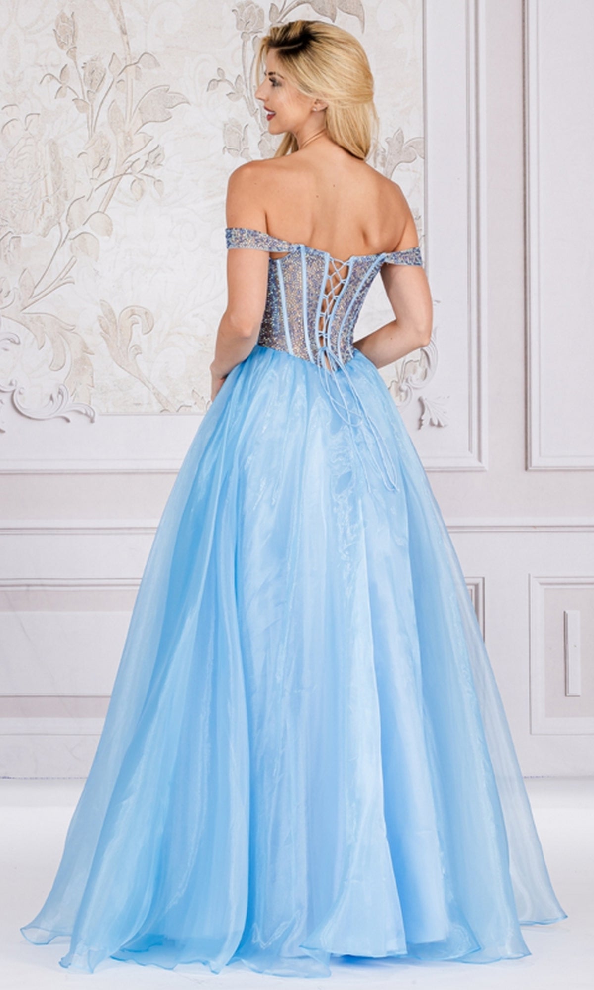 A woman stands indoors in the Organza Off-Shoulder Bodice Prom Ball Gown 7040, featuring a lace-up back and full flowing skirt, facing away to highlight the dress's intricate details.