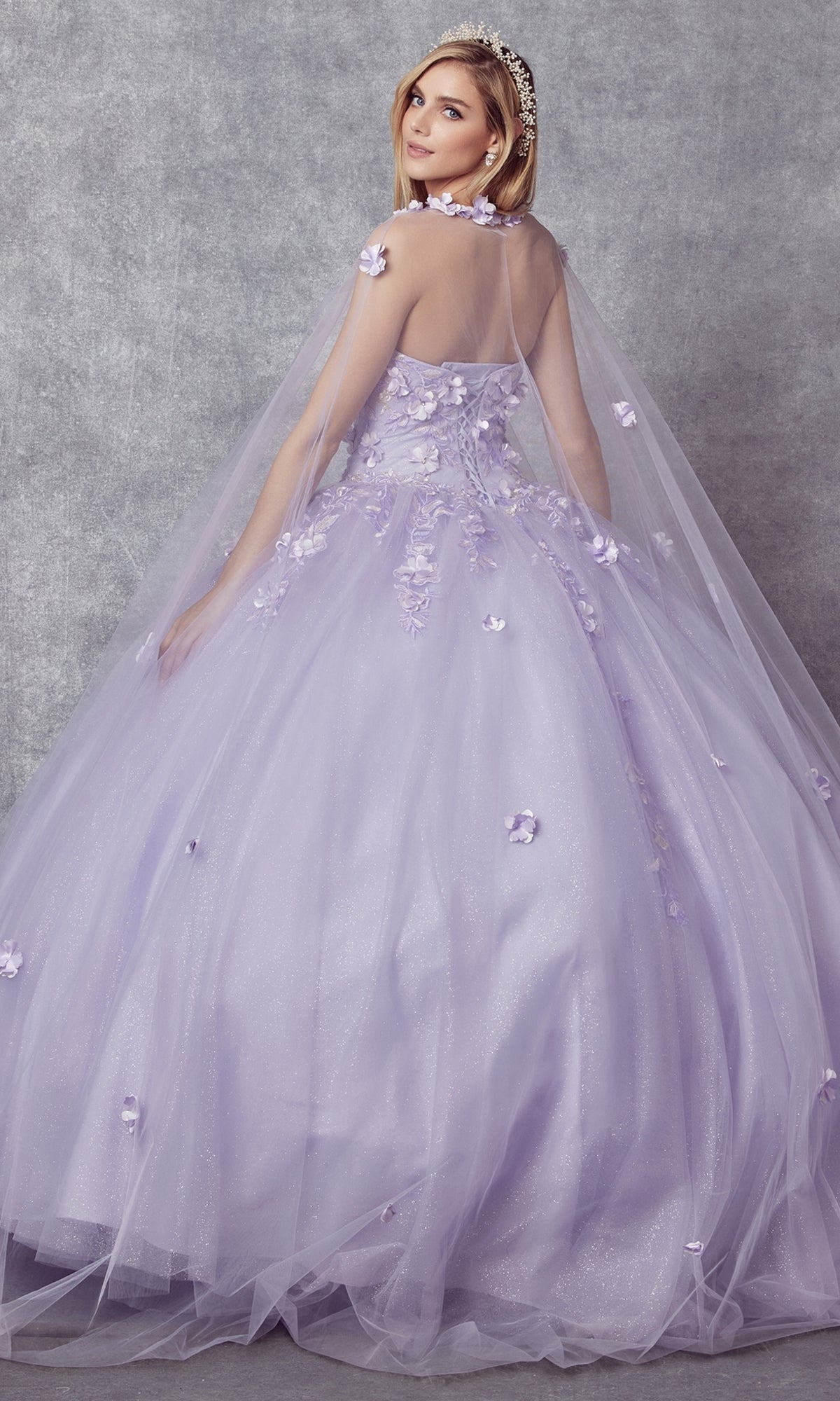 A woman in the Juliet 1435 Strapless Ball Gown Quinceañera Dress, featuring floral appliqué and a sheer cape, poses against a gray textured backdrop.