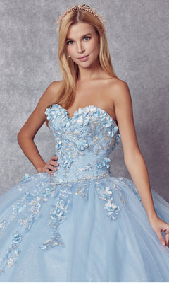 A woman wears the Juliet 1435 Strapless Ball Gown Quinceañera Dress, featuring a light blue floral embroidered bodice and tulle skirt, standing against a gray textured background.