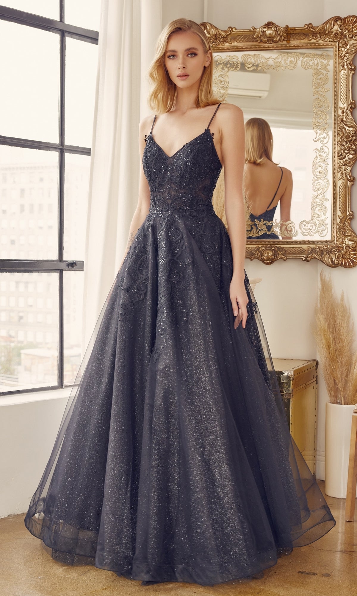 A woman stands indoors by a large window and ornate mirror, wearing the Embroidered Glitter-Tulle Long Prom Ball Gown 251 with a fitted bodice and full skirt.