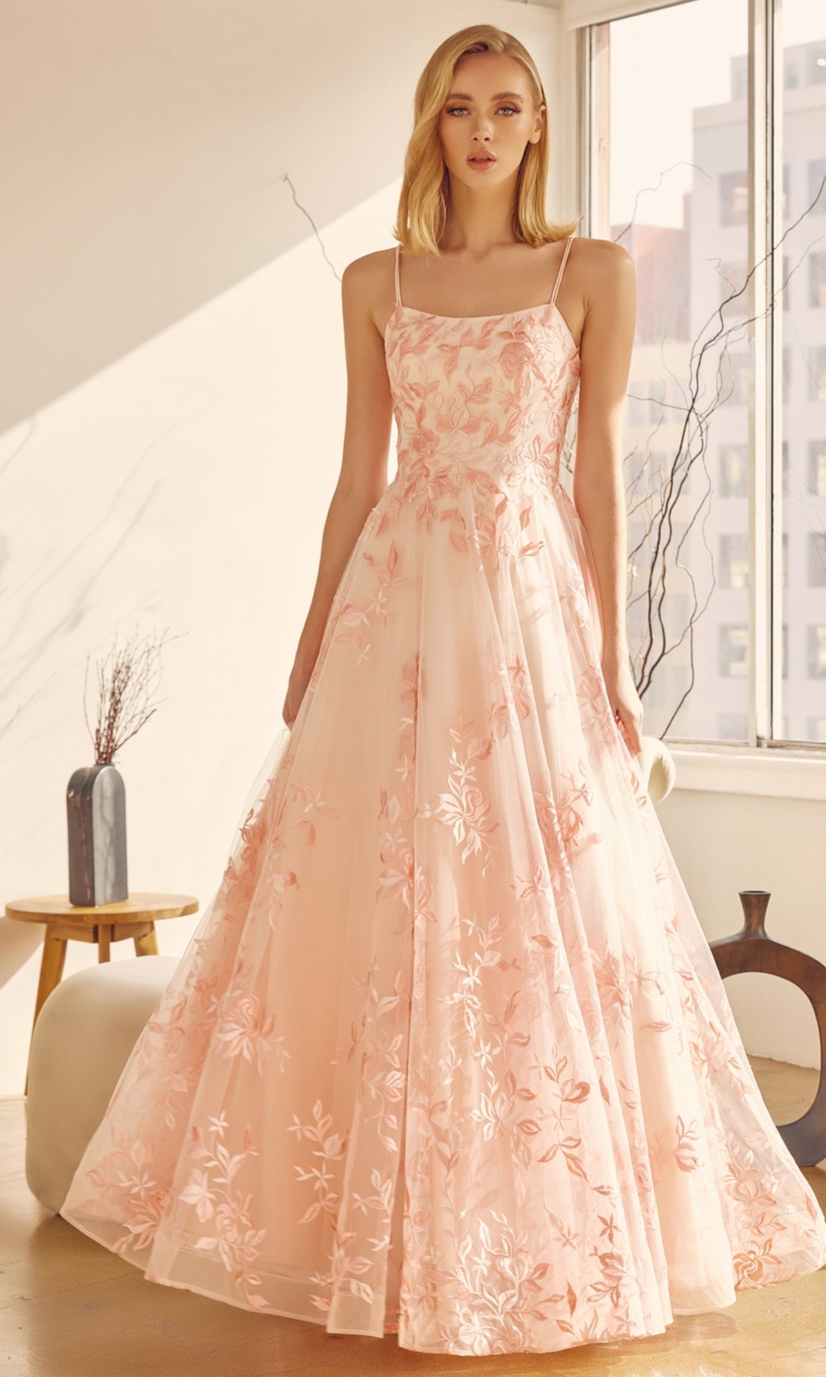 A woman stands indoors wearing the Embroidered-Lace Long A-Line Prom Ball Gown 279 in pale pink floral fabric with thin straps. Sunlight pours through large windows, illuminating the elegant setting with minimal decor.