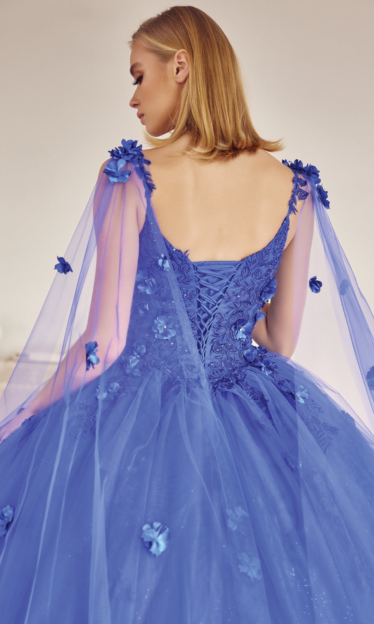 A woman stands facing away from the camera, wearing the Juliet 1436 Glitter Quinceañera Dress with Cape—a blue ball gown featuring floral appliqués and a lace-up back.