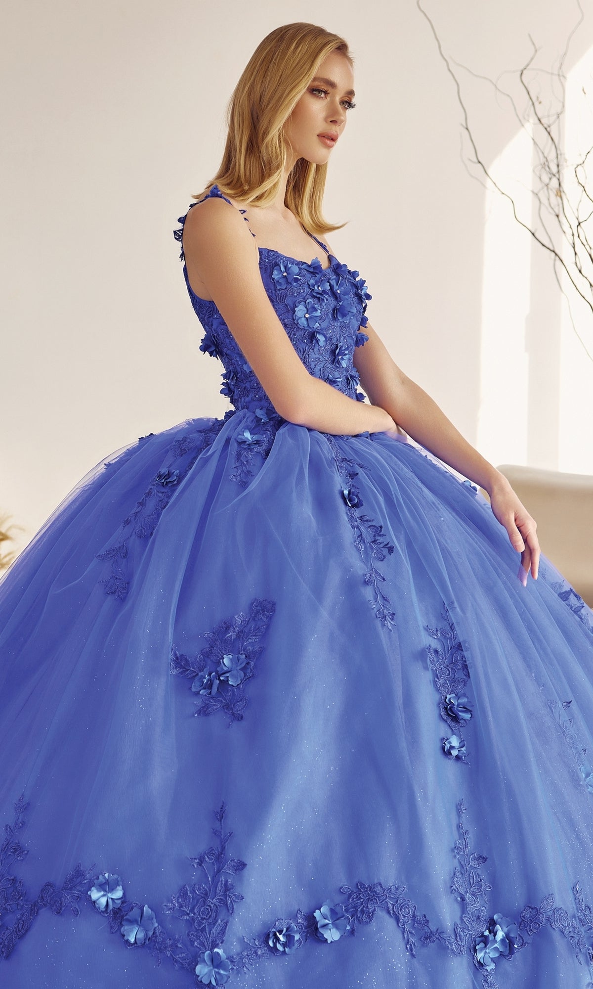 A woman wearing the Juliet 1436 Glitter Quinceañera Dress with Cape, featuring a royal blue hue, floral appliqués, and a full tulle skirt, stands indoors against a neutral backdrop.