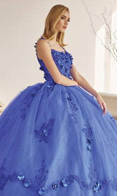 A woman wearing the Juliet 1436 Glitter Quinceañera Dress with Cape, featuring a royal blue hue, floral appliqués, and a full tulle skirt, stands indoors against a neutral backdrop.