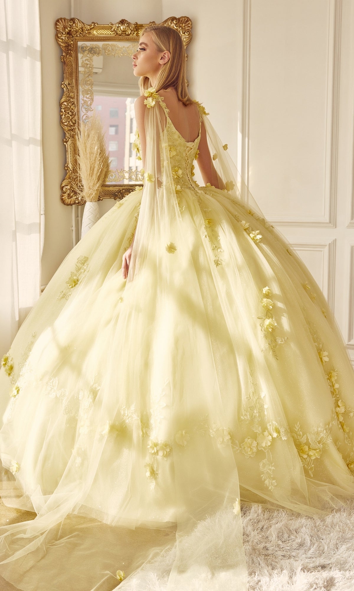 A woman in the Juliet 1436 Glitter Quinceañera Dress with Cape, featuring floral details, stands by a window with a gold-framed mirror and white walls behind her.