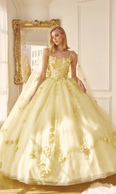 A woman stands indoors in front of a large ornate mirror wearing the Juliet 1436 Glitter Quinceañera Dress with Cape, featuring a voluminous pale yellow skirt, floral embellishments, and tulle.