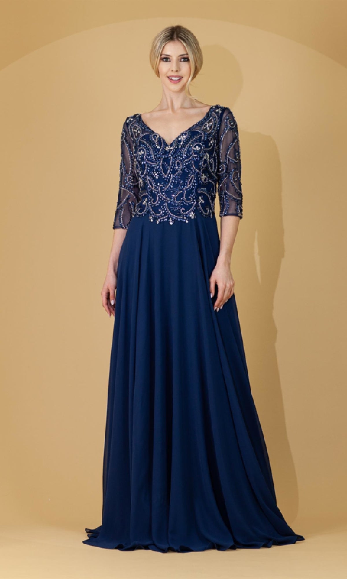 A woman poses against a beige background in the Amelia Couture 7046—a floor-length navy gown with a beaded sheer half-sleeve bodice and flowing skirt, an elegant choice for a mother of the bride.