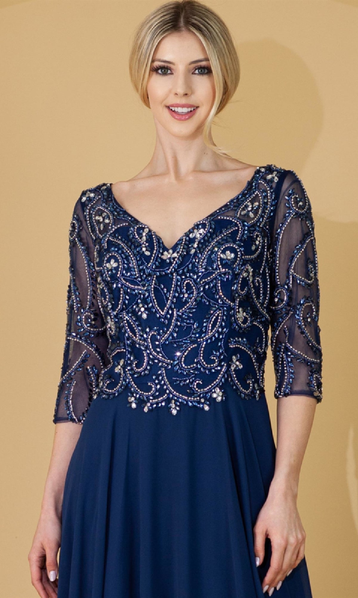 A woman smiles against a beige background, wearing the Amelia Couture 7046 sleeved long formal dress in navy blue, featuring intricate beadwork and sheer three-quarter sleeves.